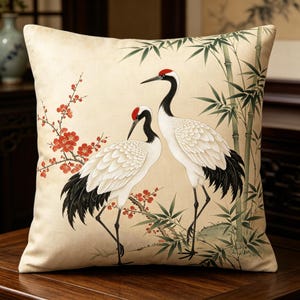 May include: A square throw pillow featuring a cream-colored background with a printed illustration of two white cranes with black wing tips and red crowns. The scene includes green bamboo stalks and a branch with small red blossoms.