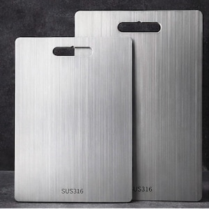 May include: Two stainless steel chopping boards with a brushed metal finish. Each board has a rectangular shape with a handle cutout. The text "SUS316" is printed on the bottom of the smaller board and "US316" on the larger board.