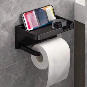 May include: Black metal toilet paper holder with a built-in shelf. A smartphone and a smartwatch rest on the shelf above a roll of white toilet paper. The holder is mounted on a gray tiled wall.