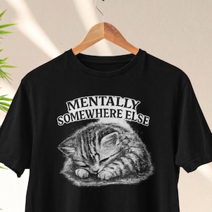 Può includere: T-shirt nero con un'illustrazione in scala di grigi di un gattino che dorme. Il testo "MENTALLY SOMEWHERE ELSE" è sopra il gattino. La maglietta è appesa a una gruccia di legno.