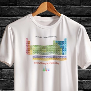 Puede incluir: Camiseta blanca con una tabla periódica de elementos colorida. La camiseta incluye el texto "Periodic Table of Elements" y "Everything is chemistry" en rojo. La camiseta está colgada en una percha de madera.