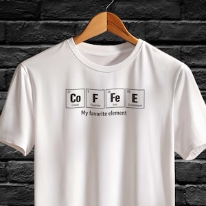 Puede incluir: Camiseta blanca con la palabra "COFFEE" escrita con los símbolos de la tabla periódica para Cobalto, Flúor, Hierro y Einstenio. Debajo está la frase "My favorite element". La camiseta está colgada de una percha de madera.