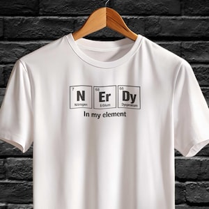Puede incluir: Camiseta blanca con la palabra "Nerdy" escrita con los símbolos de la tabla periódica de nitrógeno, erbio y disprosio. El texto "In my element" está debajo. La camiseta está colgada de una percha de madera.