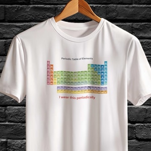 Puede incluir: Camiseta blanca con un diseño colorido de la tabla periódica. El texto "Periodic Table of Elements" está encima de la tabla, y "I wear this periodically" debajo. La camiseta está colgada en una percha de madera.