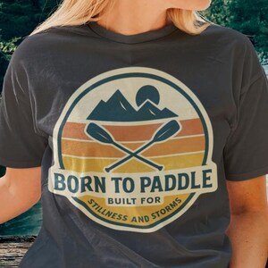 以下が含まれることがあります： 円形のグラフィックデザインが施された濃いグレーのTシャツ。デザインには、交差したパドル、山、そして「Born to Paddle」の文字が含まれています。デザインはオレンジ、黄色、青を使用し、「Built for Stillness and Storms」という言葉が添えられています。