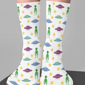 May include: White socks with a repeating pattern of green aliens, purple and blue UFOs, and yellow stars. The socks are knee-high and have a playful, whimsical design.