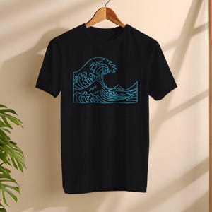 Puede incluir: Camiseta negra con un diseño de ola azul. El gráfico representa una ola oceánica estilizada con crestas y una montaña en el fondo. La camiseta está colgada en una percha de madera.