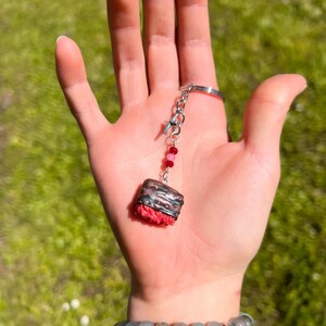 May include: A handmade keychain featuring a miniature sushi roll charm. The charm has a red, textured base, a silver-colored band, and a dark top. It hangs from a silver chain with pink and red beads.