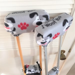 May include: Gray fleece crutch covers with a repeating pattern of cartoon dogs, paw prints, and the text "I Woof You". The covers are designed to fit the arm and hand rests of crutches, providing comfort and style.