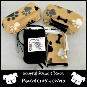 May include: Three beige padded crutch covers with a paw print and bone design. The covers are made of soft fleece and have a black velcro strap. A black pouch with the text "Clutch 'Crutch' Purse My-S-Cap" is also shown. The text on the pouch reads: "Perfect for those small can't do without essentials. Attaches to hand grip on a crutch. Velcro closes. Machine Washable. Tumble Dry. Polyester. Made in Kentucky. www.mysyscap.etsy.com"