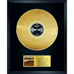 May include: Framed gold record of the System of a Down album "Toxicity." The record is centered on a black background within a black frame. A small plaque below the record provides album details. The record is a collectible item.