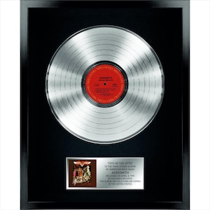 Peut inclure: Un disque de platine encadré pour l'album Toys in the Attic d'Aerosmith. L'objet présente un disque vinyle argenté avec une étiquette centrale rouge, monté sur un fond noir avec une plaque argentée détaillant les informations de l'album.