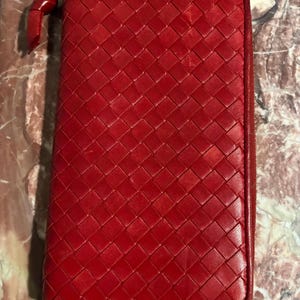 May include: A red, rectangular wallet with a woven pattern. The wallet has a zipper closure and a small loop on the zipper pull. The overall design is a classic and elegant style.
