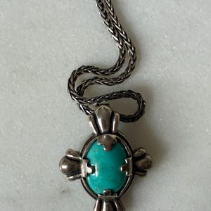 May include: Silver-toned pendant necklace featuring a cross design with a turquoise-colored oval stone at the center. The necklace has a braided chain. The cross has decorative elements on each arm.