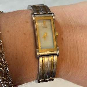 May include: A rectangular gold and silver-toned watch with a gold-colored key-shaped design on the face. The watch band is a combination of silver and gold-toned metal. The watch is on a wrist.