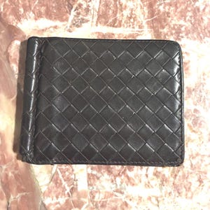 May include: A dark brown leather money clip wallet with a woven pattern. The wallet has a metal clip on the left side and is rectangular in shape. The leather has a matte finish.