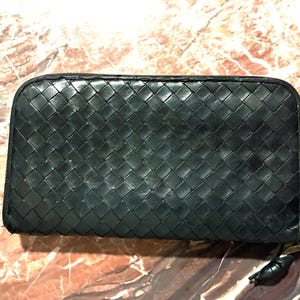 May include: A black, rectangular wallet with a woven leather design. The wallet has a zipper closure and a small, black leather pull. The wallet is on a marble-patterned surface.