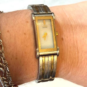 May include: A rectangular gold and silver-toned wristwatch with a white face and gold hands. The watch band is a combination of silver and gold links. The watch is on a wrist with a silver chain bracelet.