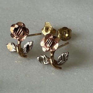 May include: A pair of rose-shaped stud earrings. The rose petals are a warm rose gold color, with gold stems and silver leaves. The earring backs are gold. The earrings are displayed on a light gray surface.
