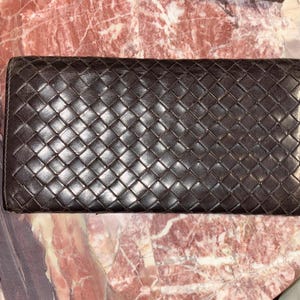 May include: A dark brown leather wallet with a woven design. The rectangular wallet is closed. The surface has a subtle sheen, suggesting a polished finish. The wallet is likely designed for carrying cards and cash.