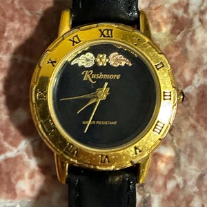 Puede incluir: Un reloj de pulsera Rushmore dorado con esfera negra y agujas doradas. El reloj presenta números romanos y una correa de cuero negro. La esfera del reloj tiene la inscripción "WATER RESISTANT".