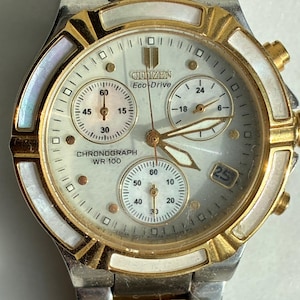 May include: A Citizen Eco-Drive chronograph watch with a silver and gold-tone stainless steel band. The watch face is white with gold accents, three sub-dials, and a date display. The watch has a WR 100 rating.