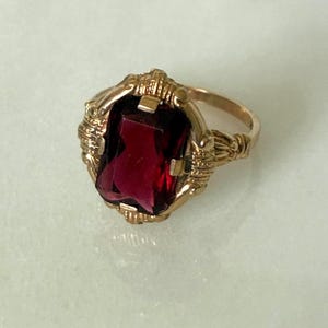 May include: A gold ring with a large, rectangular, deep red gemstone. The ring has an ornate, textured setting with detailed filigree work around the gemstone. The band is smooth and polished.