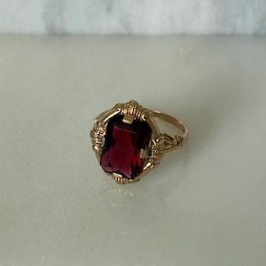 May include: A gold-toned ring featuring a large, rectangular, deep red gemstone. The ring has an ornate, detailed setting with decorative elements around the gemstone. The band is smooth and simple, contrasting with the elaborate setting.