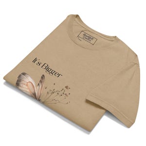May include: Tan t-shirt with the text "It's Bigger" and a butterfly graphic. The butterfly has brown and beige wings. The shirt also has a tag that says "Heartfelt Quotes".