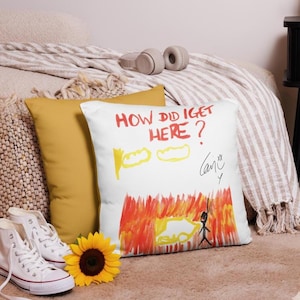 May include: White square pillow with a colorful drawing and the text "HOW DID I GET HERE?". The drawing features yellow clouds, red flames, and a stick figure. A yellow pillow and a sunflower are also visible.