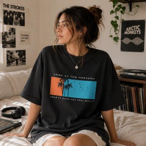 K&ouml;nnte beinhalten: Ein anthrazitfarbenes Oversize-T-Shirt mit einer rechteckigen Grafik auf sonnenuntergangsorangem und himmelblauem Hintergrund. Die Grafik zeigt schwarze Silhouetten von Palmen und V&ouml;geln sowie den Text LOOK AT THE HORIZON DOES IT MAKE YOU FEEL SMALL?