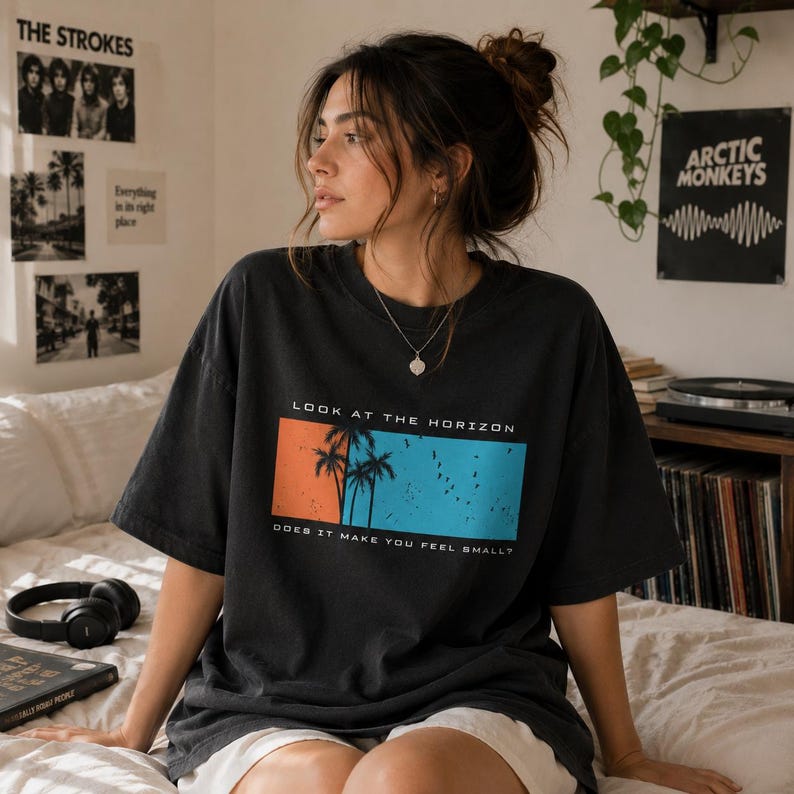 K&ouml;nnte beinhalten: Ein anthrazitfarbenes Oversize-T-Shirt mit einer rechteckigen Grafik auf sonnenuntergangsorangem und himmelblauem Hintergrund. Die Grafik zeigt schwarze Silhouetten von Palmen und V&ouml;geln sowie den Text LOOK AT THE HORIZON DOES IT MAKE YOU FEEL SMALL?