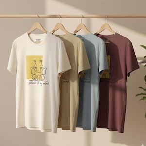 以下が含まれることがあります： 木製の棒に、クリーム色、ベージュ色、水色、バーガンディ色の半袖Tシャツが4枚吊るされています。各シャツには、城の線画と黒い文字で「palaces of my mind」が描かれた黄色の正方形があります。