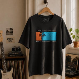 K&ouml;nnte beinhalten: Ein anthrazitfarbenes Kurzarm-T-Shirt mit einer rechteckigen Grafik, die Palmensilhouetten vor einem orange-blauen Hintergrund zeigt. Der Text "LOOK AT THE HORIZON" steht &uuml;ber der Grafik und "DOES IT MAKE YOU FEEL SMALL?" darunter.