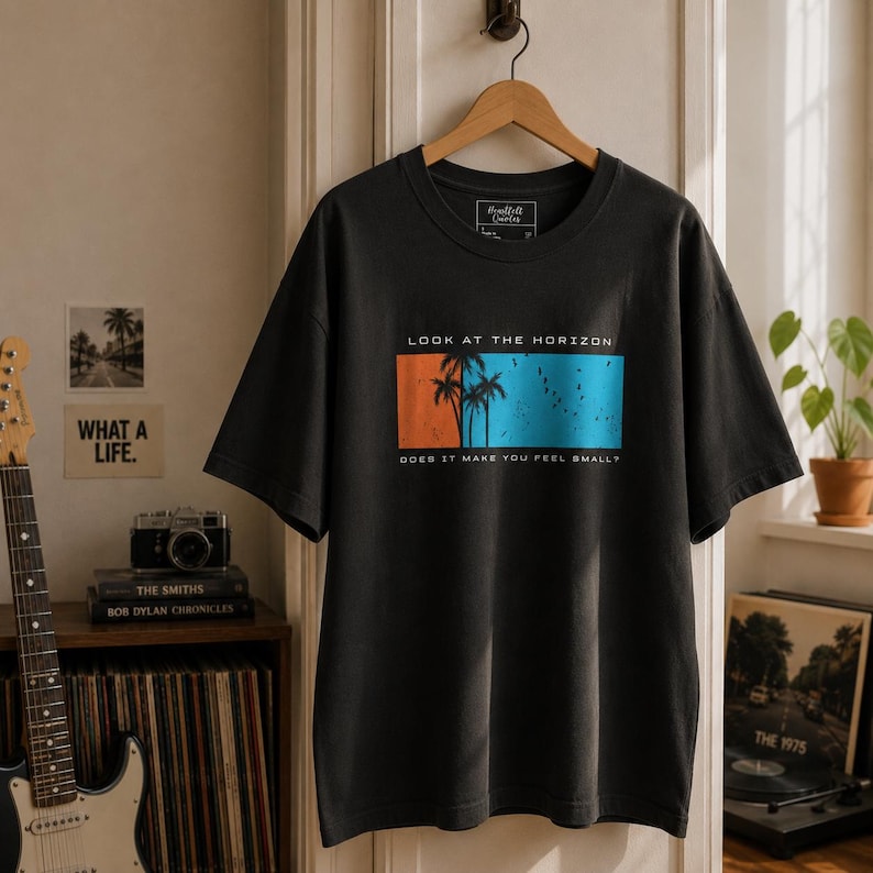 K&ouml;nnte beinhalten: Ein anthrazitfarbenes Kurzarm-T-Shirt mit einer rechteckigen Grafik, die Palmensilhouetten vor einem orange-blauen Hintergrund zeigt. Der Text "LOOK AT THE HORIZON" steht &uuml;ber der Grafik und "DOES IT MAKE YOU FEEL SMALL?" darunter.
