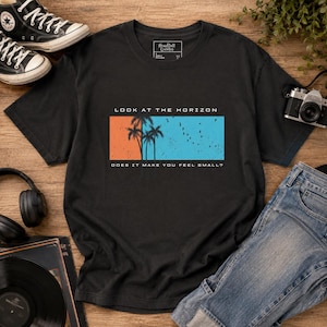 K&ouml;nnte beinhalten: Ein schwarzes Kurzarm-T-Shirt mit einer rechteckigen Grafik, die eine orangefarbene linke Seite und eine blaue rechte Seite aufweist. Das Motiv zeigt Palmensilhouetten, V&ouml;gel sowie den Text "LOOK AT THE HORIZON" und "DOES IT MAKE YOU FEEL SMALL?"