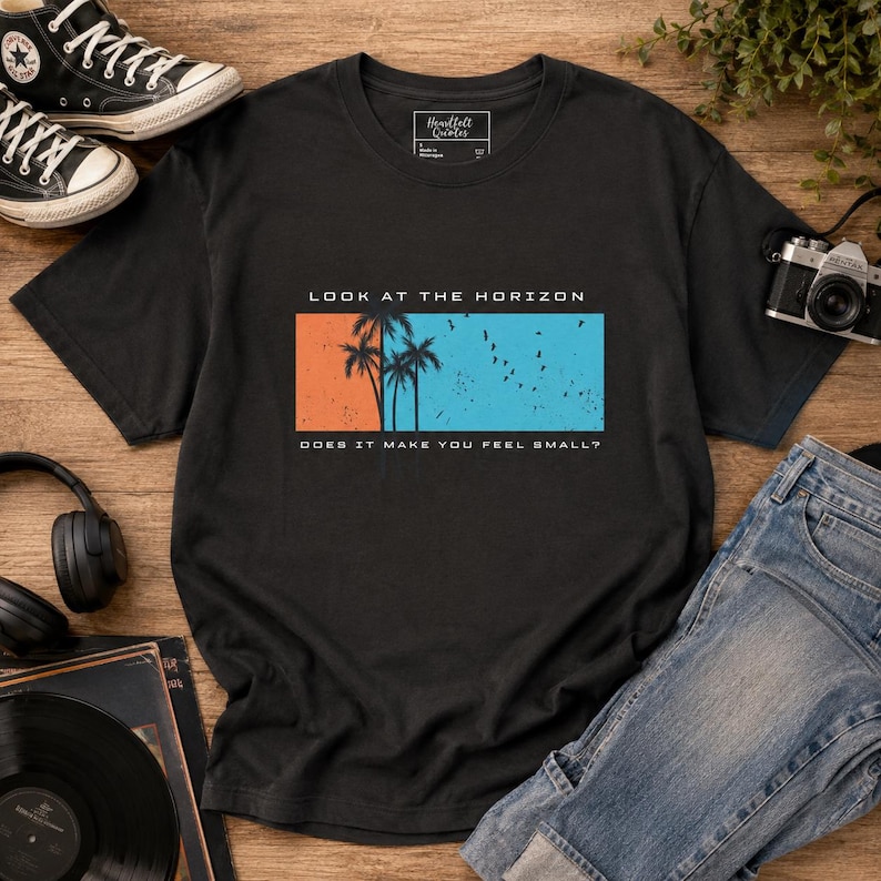 K&ouml;nnte beinhalten: Ein schwarzes Kurzarm-T-Shirt mit einer rechteckigen Grafik, die eine orangefarbene linke Seite und eine blaue rechte Seite aufweist. Das Motiv zeigt Palmensilhouetten, V&ouml;gel sowie den Text "LOOK AT THE HORIZON" und "DOES IT MAKE YOU FEEL SMALL?"