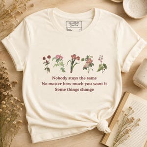 Puede incluir: Una camiseta de manga corta color crema con una fila de cinco ilustraciones botánicas de flores en tonos suaves. Debajo de las flores, el texto dice: Nobody stays the same, No matter how much you want it, Some things change.