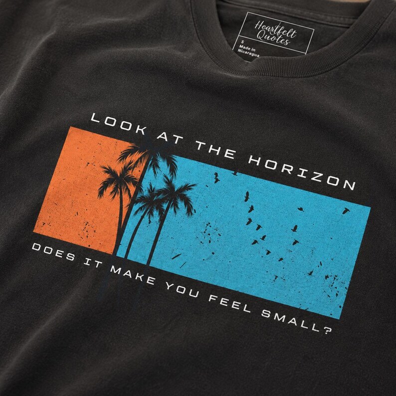 K&ouml;nnte beinhalten: Ein anthrazitfarbenes T-Shirt mit einer rechteckigen Grafik auf orangefarbenem und blauem Hintergrund. Das Motiv zeigt Silhouetten von Palmen und V&ouml;geln. Der Text lautet: LOOK AT THE HORIZON DOES IT MAKE YOU FEEL SMALL? Ein Heartfelt Quotes Etikett ist sichtbar.