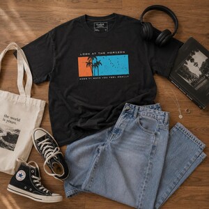 K&ouml;nnte beinhalten: Ein schwarzes T-Shirt mit einem Grafikdruck, der Palmen vor einem orange-blauen rechteckigen Hintergrund zeigt, mit dem Text LOOK AT THE HORIZON und DOES IT MAKE YOU FEEL SMALL? Eine hellblaue Jeans, eine Stofftasche und Turnschuhe.