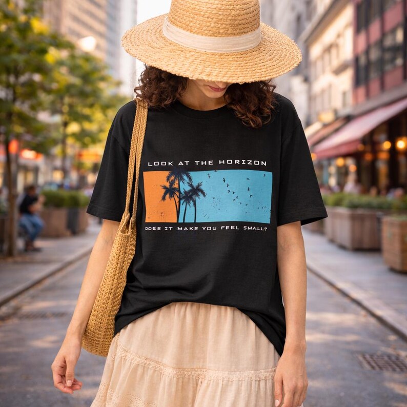 K&ouml;nnte beinhalten: Ein schwarzes T-Shirt mit einer rechteckigen Grafik, die einen orange-blauen Sonnenuntergang, Palmensilhouetten und V&ouml;gel zeigt. Der Text lautet LOOK AT THE HORIZON und DOES IT MAKE YOU FEEL SMALL? Kombiniert mit einem Strohhut und einer geflochtenen Tasche.