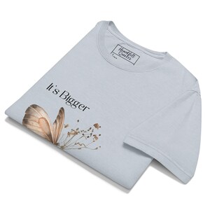 May include: Light blue t-shirt with a butterfly and floral design. The words "It's Bigger" are printed on the shirt. The shirt has a tag that says "Heartfelt Quotes".