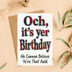 May include: A white greeting card featuring the text "Och, it's yer Birthday" in a red, green, and blue plaid pattern with a silver outline, followed by the black text "Ah Cannae Believe Ye're That Auld" printed on the lower portion of the card.