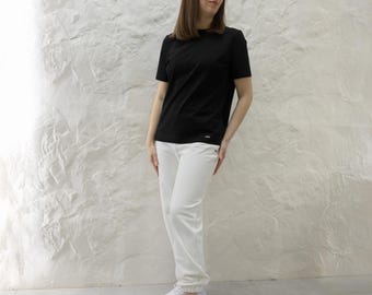 May include: A woman wearing a black short-sleeved t-shirt and white pants. She is standing in front of a white wall.