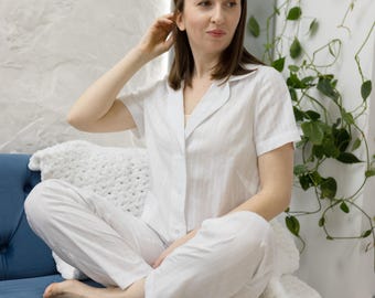 May include: A woman wearing a white button-down pajama top and matching pants sits on a blue couch with a white throw blanket. She is looking to the right of the frame.