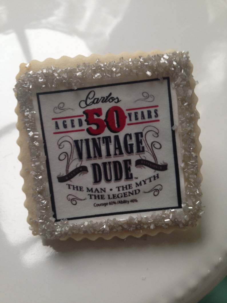 K&ouml;nnte beinhalten: Ein dekorierter Zuckerkeks mit silbernen Streuseln an den R&auml;ndern. Der Keks hat ein bedrucktes Design mit dem Text "Carlos Aged 50 Years Vintage Dude Experience The Man The Myth The Legend Courage 60% Ability 40%"