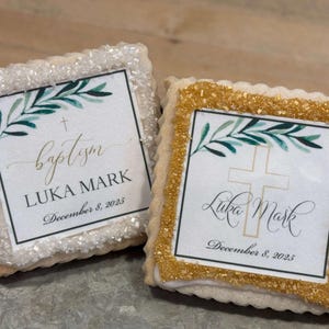 May include: Two square sugar cookies decorated with white and gold sprinkles. The cookies have a white background with a green leafy branch, a gold cross, and the text "baptism LUKA MARK December 8, 2025".