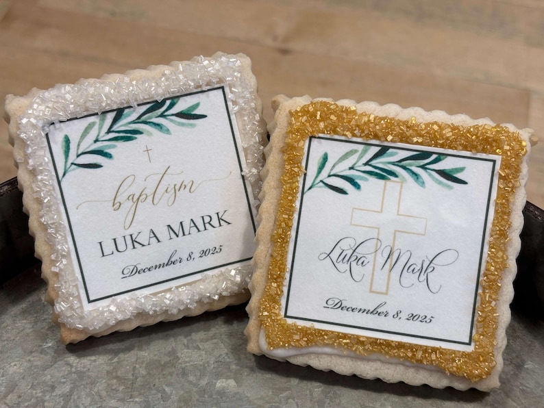 May include: Two square sugar cookies decorated with white and gold sprinkles. The cookies have a white background with a green leafy branch, a gold cross, and the text "baptism LUKA MARK December 8, 2025".