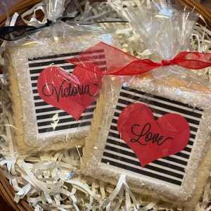 May include: Two square sugar cookies decorated with black and white stripes and red hearts. One cookie says "Victoria" inside the heart and the other says "Love".