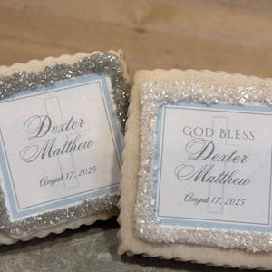 May include: Two decorated sugar cookies with white frosting and silver sprinkles. The cookies have a blue and white design with a cross and the text "God Bless Dexter Matthew August 17, 2025".